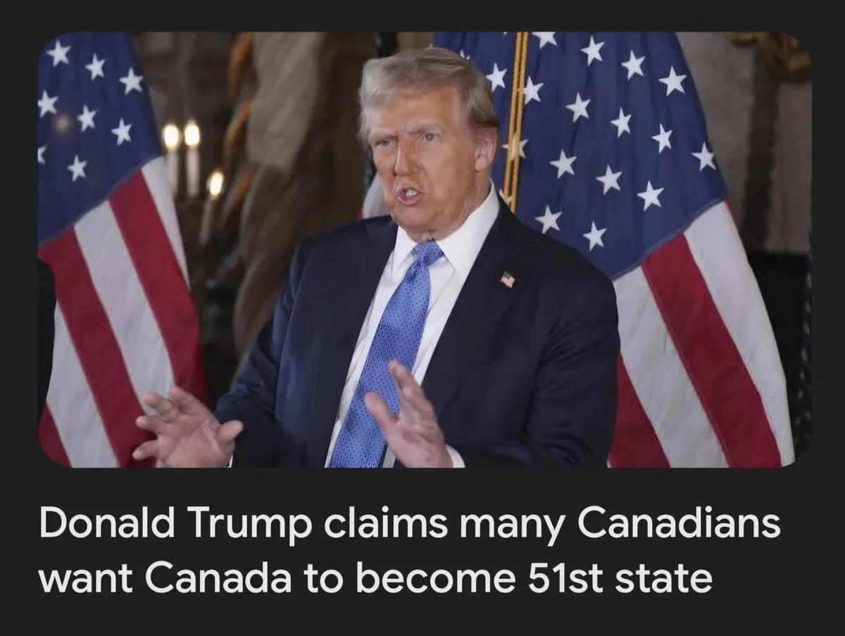 A man in a dark suit and blue tie stands in front of two American flags, gesturing with both hands as if speaking…