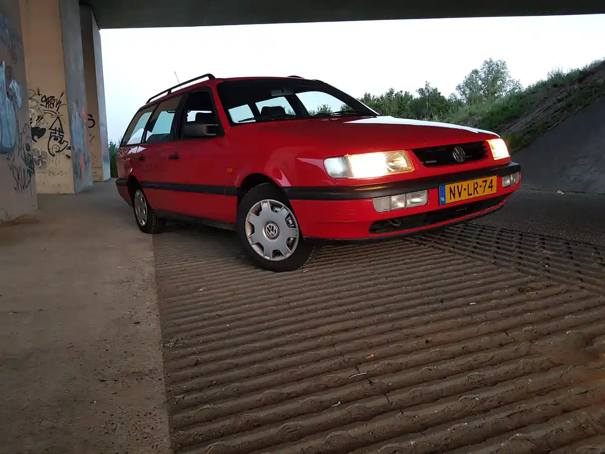 A red Volkswagen Passat station wagon is parked under a concrete overpass on a ribbed metal grate. The car's headlights are…