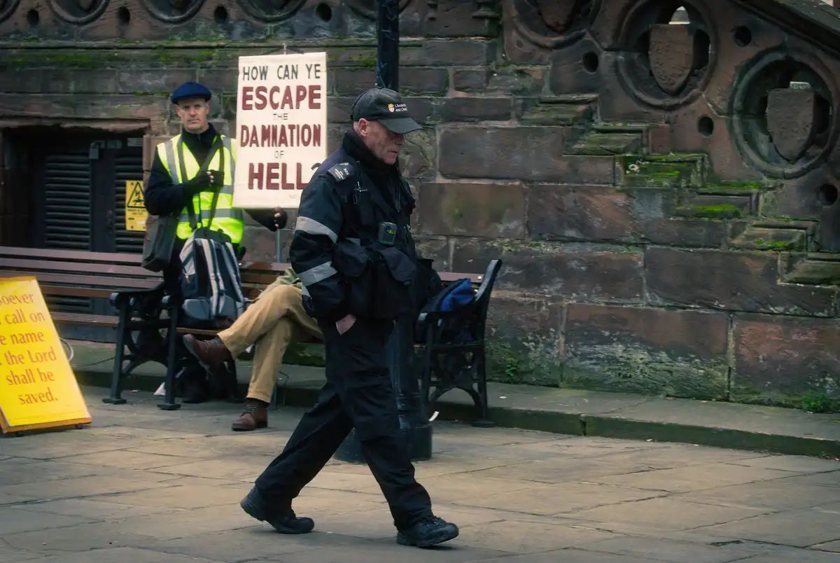 A police officer walks past a man holding a sign that reads 'HOW CAN YE ESCAPE THE DAMNATION OF HELL' while another sign…