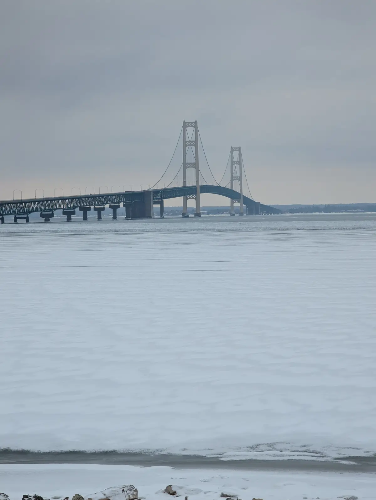 A wide shot of a large suspension bridge spanning a frozen body of water under an overcast sky. The foreground shows…