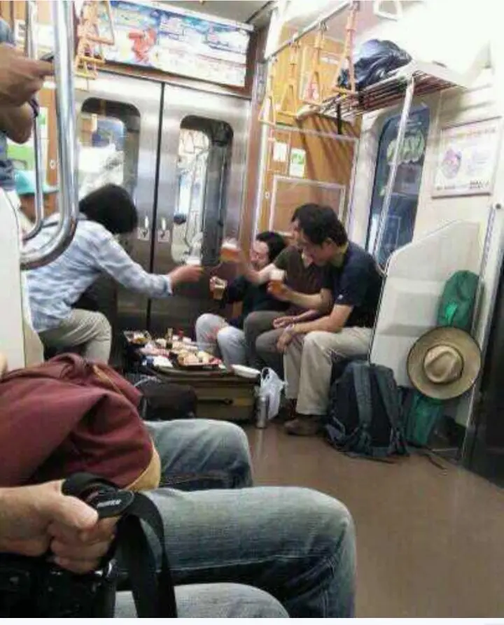Inside a crowded subway car, several people are seated around an open suitcase filled with food and drinks, raising glasses…
