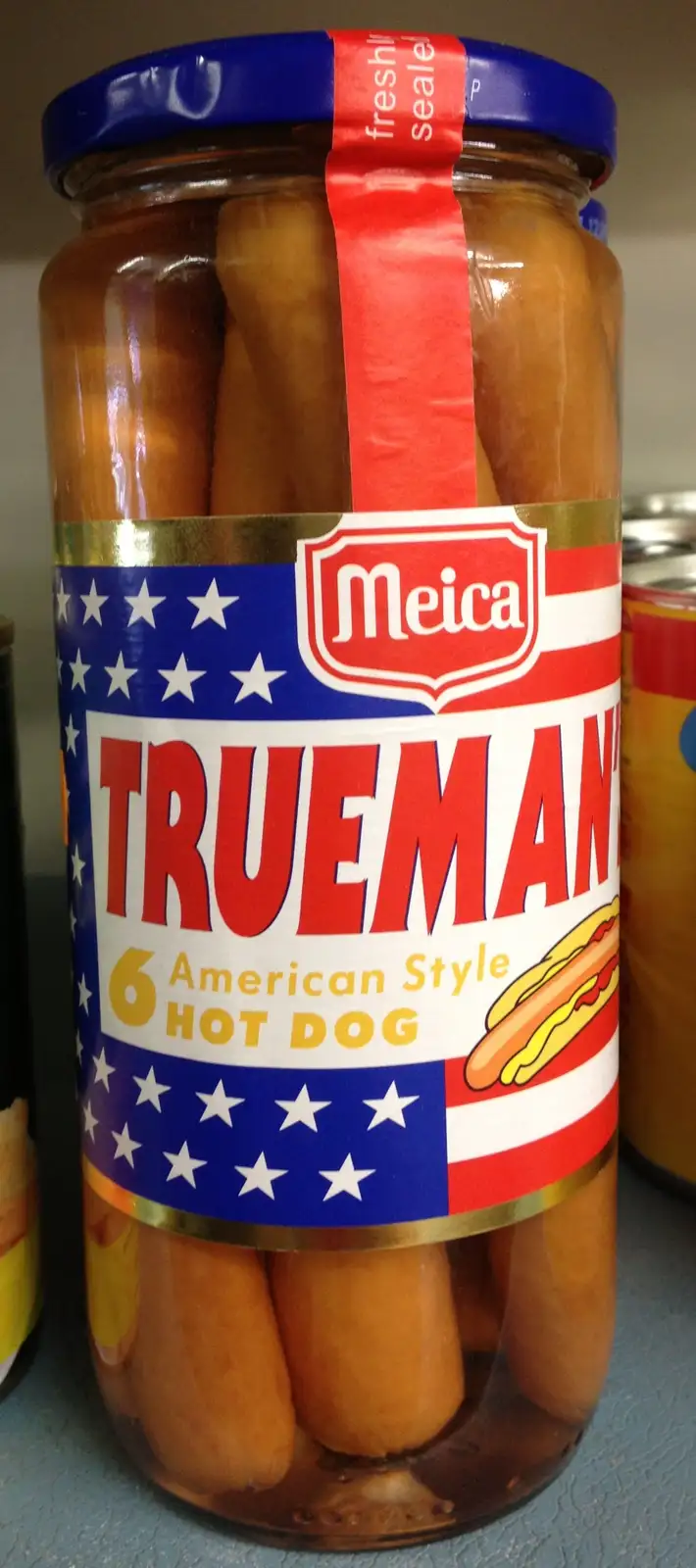 A jar of Meica brand hot dogs labeled 'TRUEMAN' with an American flag design, containing six American style hot dogs. The…