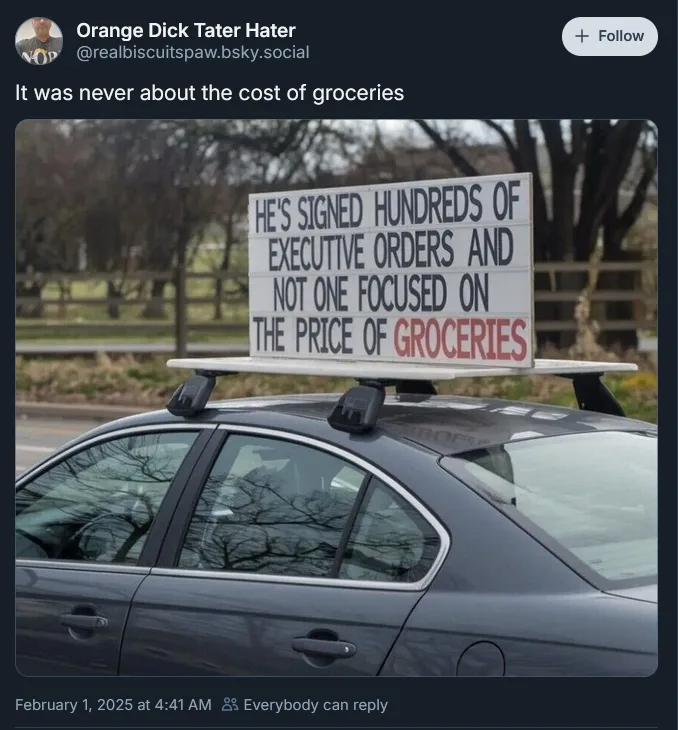 A dark gray sedan has a large white sign mounted on its roof rack, displaying text in black and red letters. The sign reads…