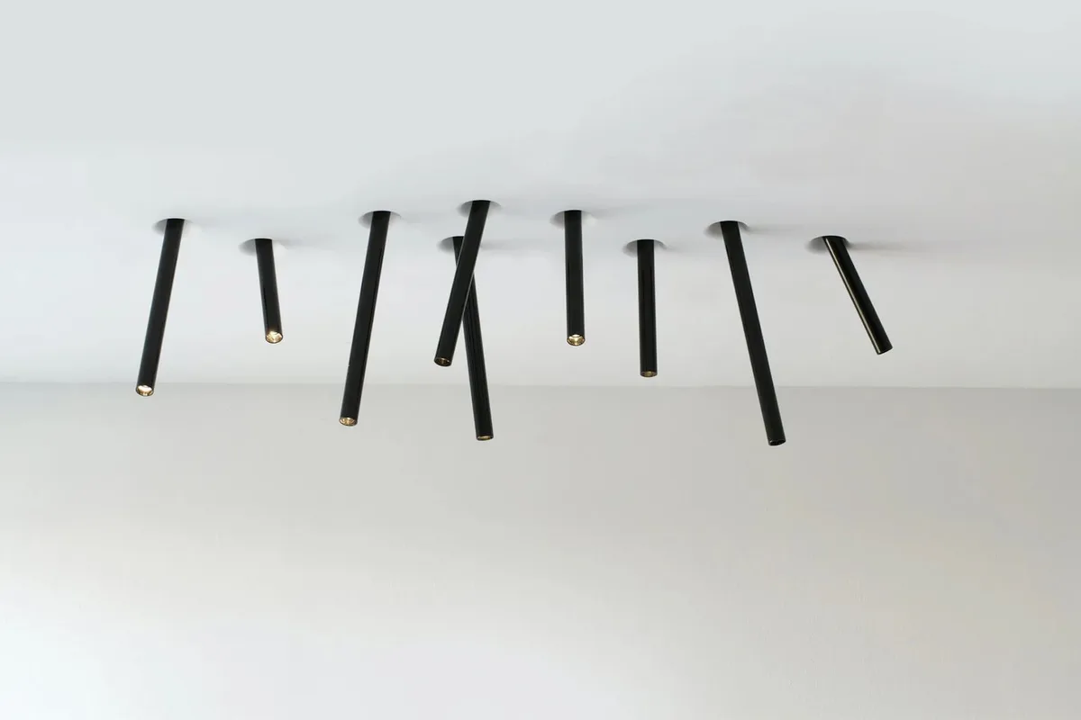 A minimalist photo shows several black cylindrical lights hanging from a white ceiling at various angles, with gold-colored…