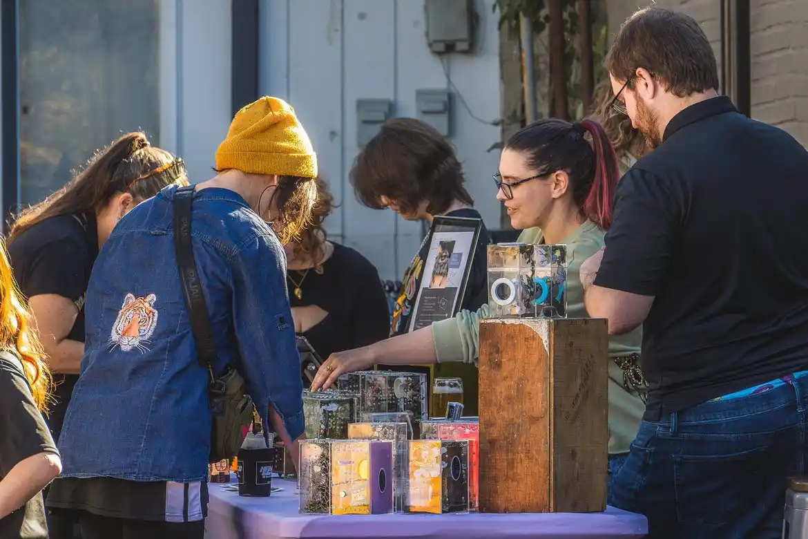 A group of people browse handmade cassette tapes at an outdoor market stall. One person in a denim jacket with a tiger patch…