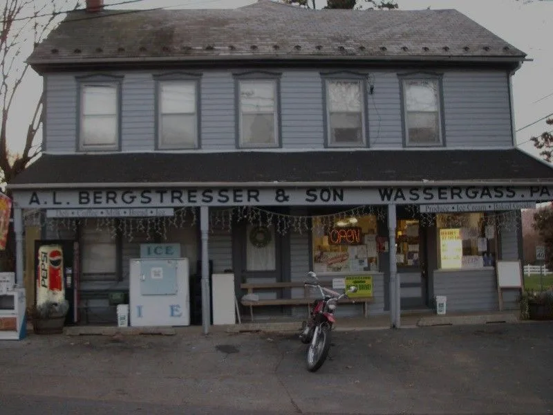 A faded photo of a two-story gray clapboard building with a storefront sign reading 'A.L. BERGSTRESSER & SON WASSERGASS.PA'…