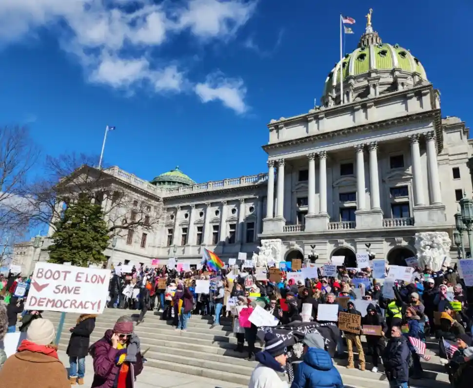 A large crowd of protesters gathers on the steps of a grand neoclassical state capitol building under a bright blue sky…