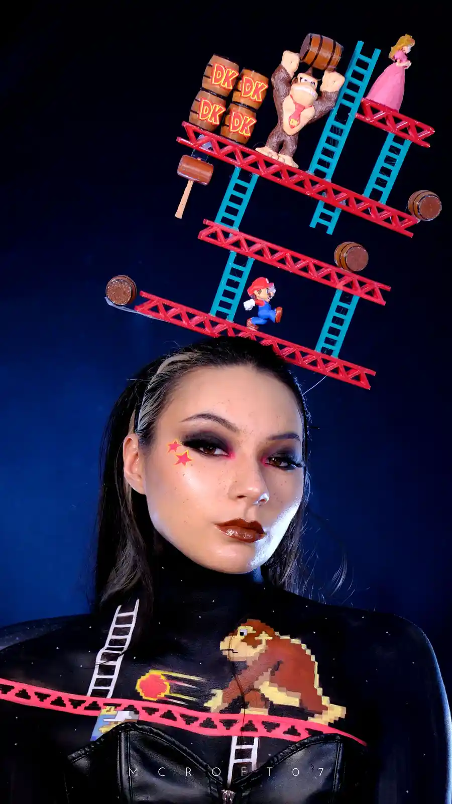 A person with dark hair and dramatic makeup, including red stars on their cheek, has a miniature Donkey Kong game set up on…