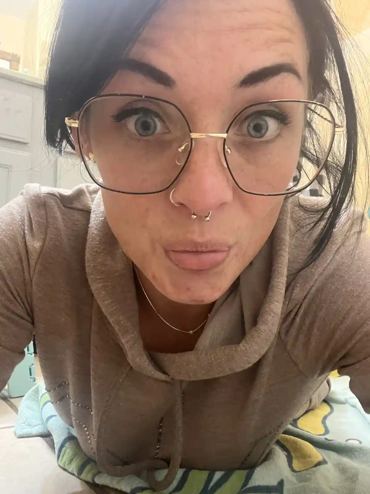 A person with dark hair, large glasses, and a nose ring stares intensely at the camera with wide eyes and a slightly pouty…