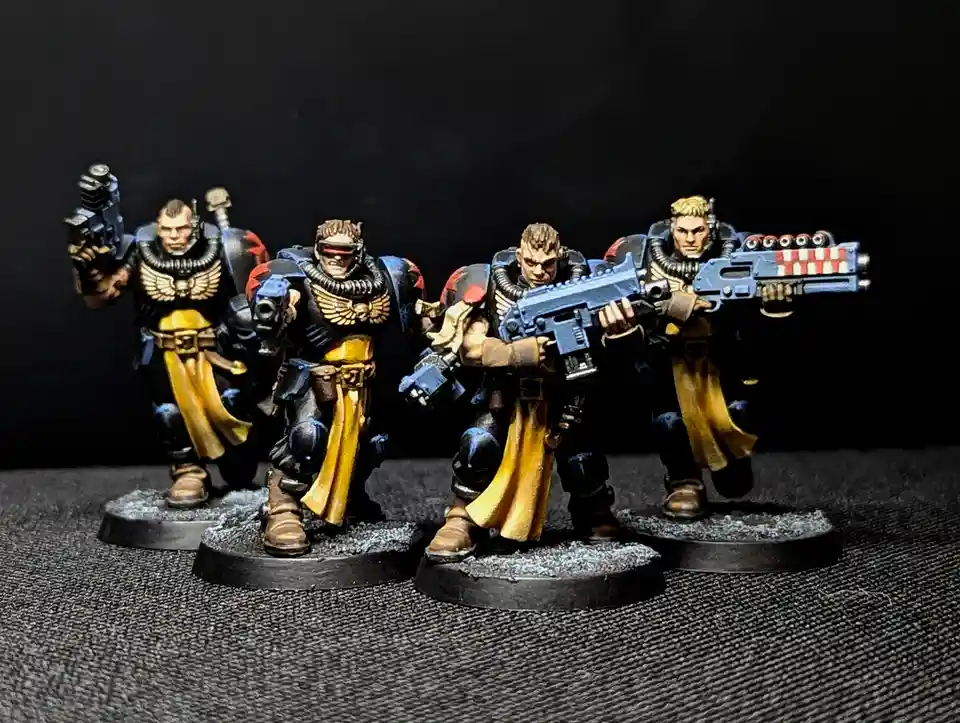 Four painted miniature figurines of space marines in black and gold armor, each holding futuristic weapons, stand on…