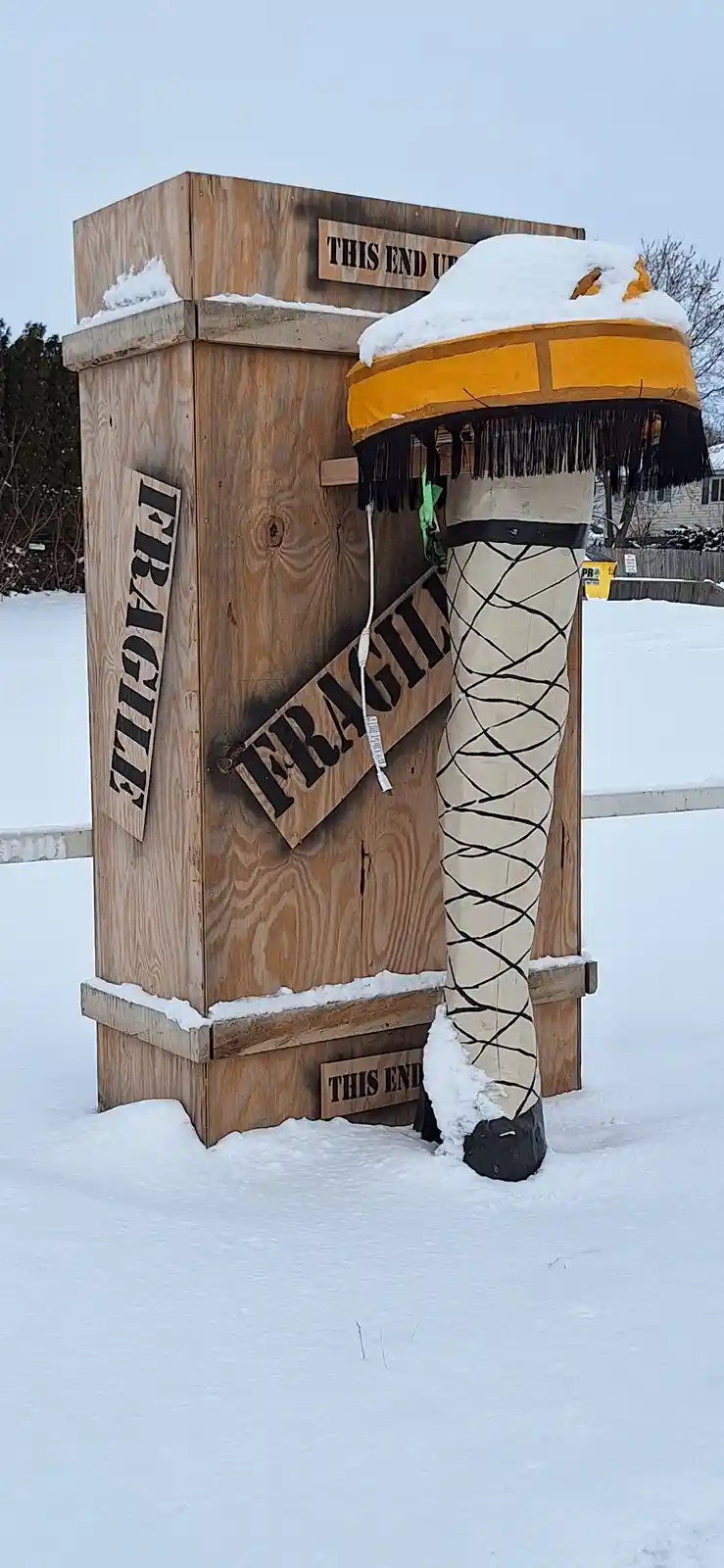 A large wooden crate labeled 'FRAGILE' and 'THIS END UP' stands in a snowy yard, with a giant, snow-capped, fringed ice…