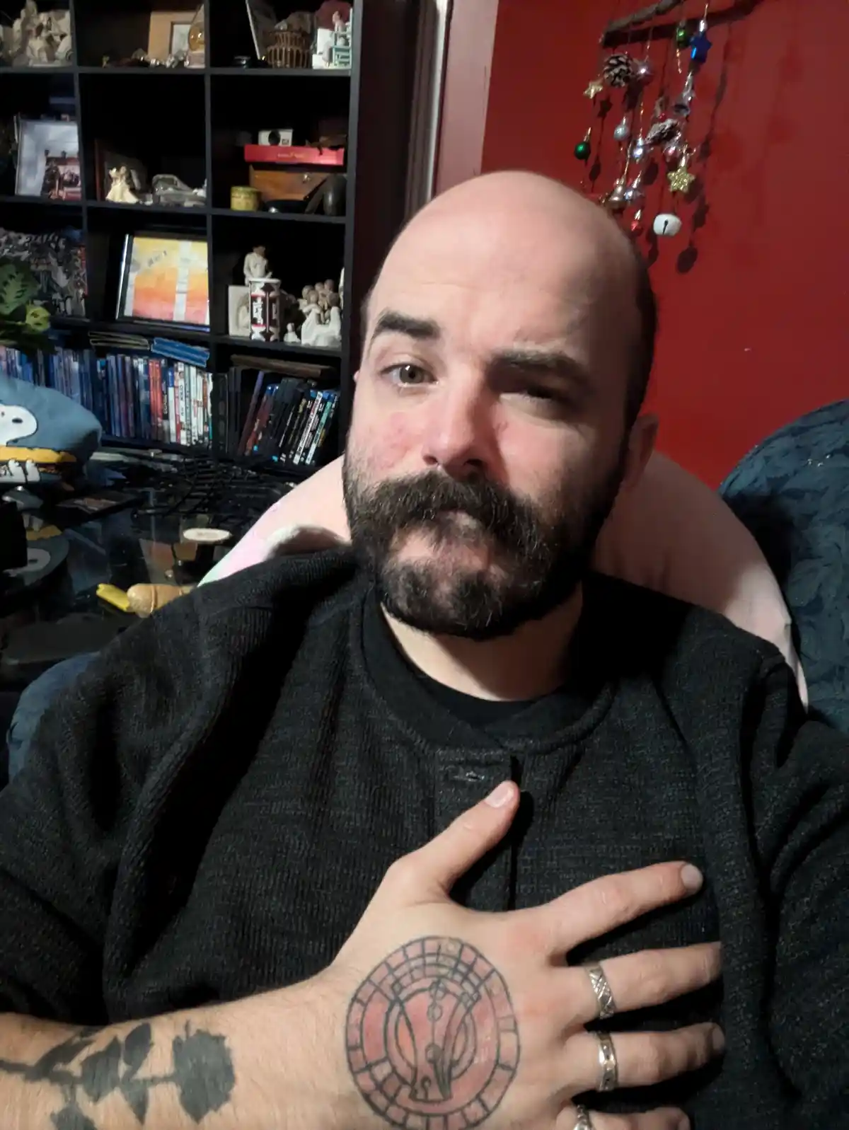 A bald man with a beard and tattoos, including a circular design on his left hand, sits in a chair with his hand over his…