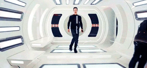 A man in a black shirt and pants stands in a futuristic white corridor with circular walls and glowing lights. He is shown…