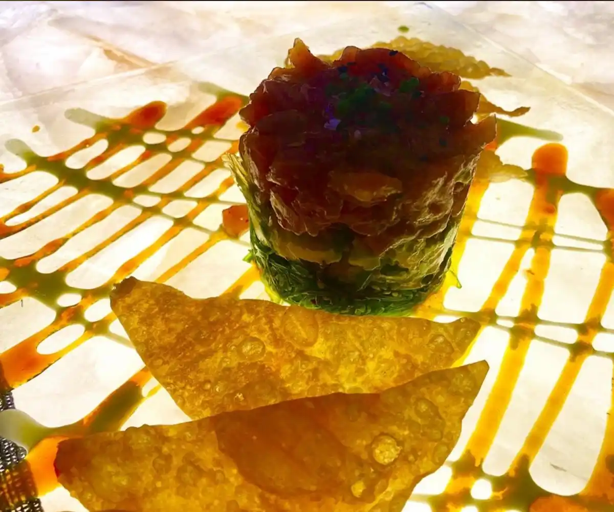 A close-up photo of an elaborately plated dish featuring a cylindrical stack of thinly sliced raw fish atop a green base…