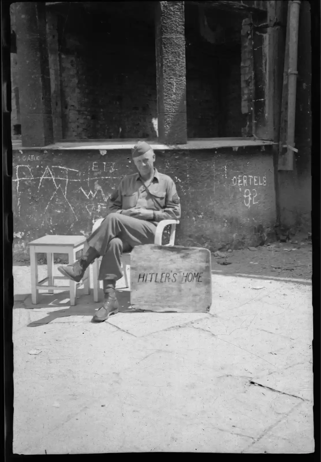 A black-and-white historical photo shows a smiling U.S. Army soldier sitting on a white plastic chair in front of a ruined…