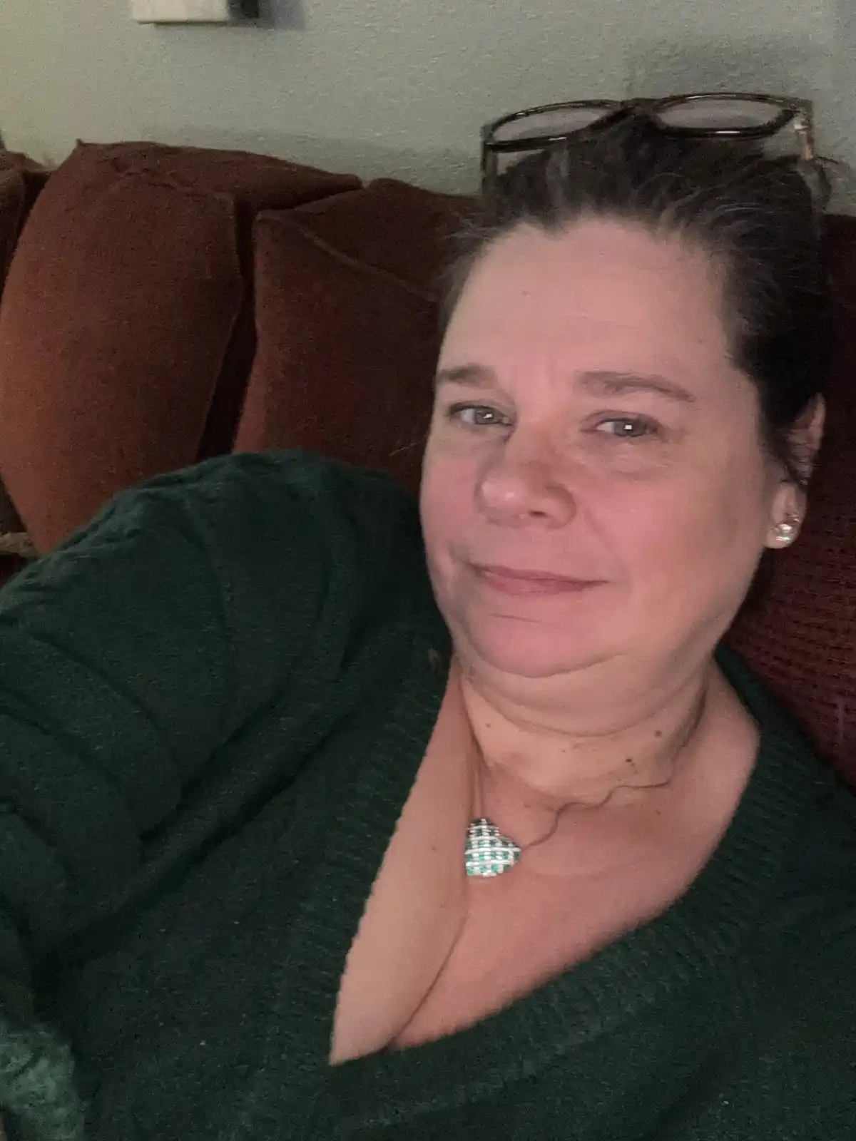 A woman with glasses perched on her head sits on a red couch, wearing a dark green sweater and a green-and-white checkered…