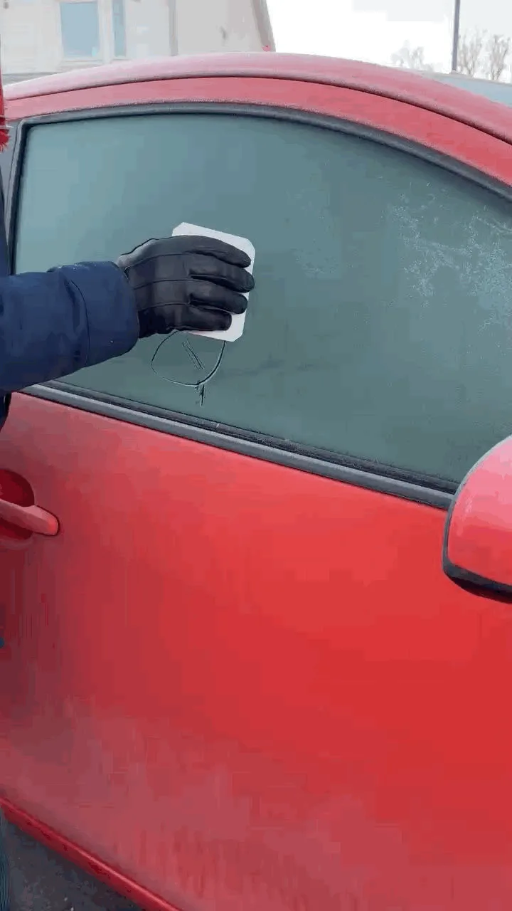 A person wearing black gloves uses a white cloth to wipe frost off a red car's side window. As they wipe, the glass shatters…