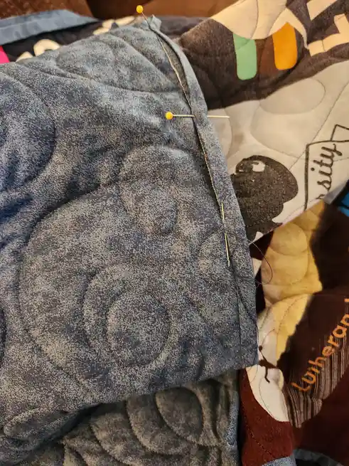 A close-up photo shows a blue denim quilt with circular stitching patterns, held together by yellow-headed pins. A needle…
