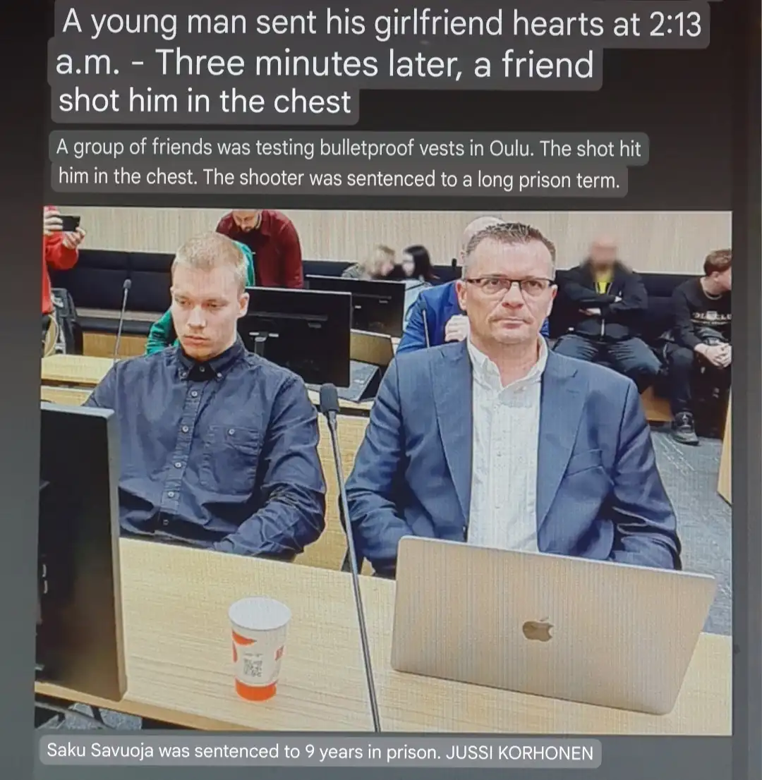 The image shows two men seated at a wooden table in what appears to be a courtroom or hearing room, with microphones and…