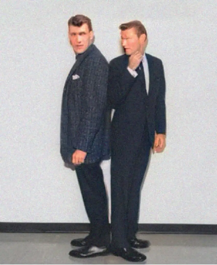 Two men in suits stand back-to-back against a plain wall; the man on the right, facing left, has his hand on his neck and…