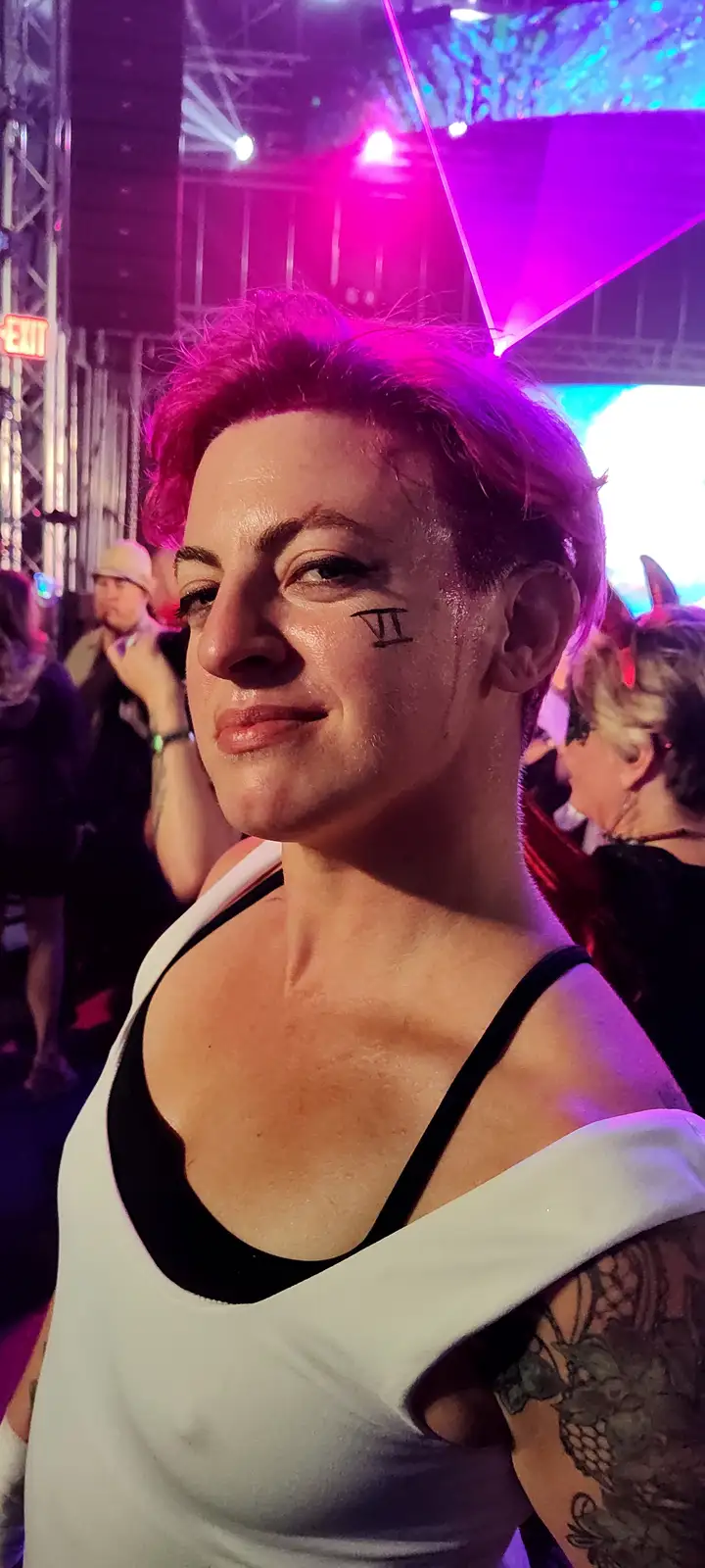 A person with short hair and a white tank top over a black bra stands in a crowded, brightly lit venue with purple and blue…