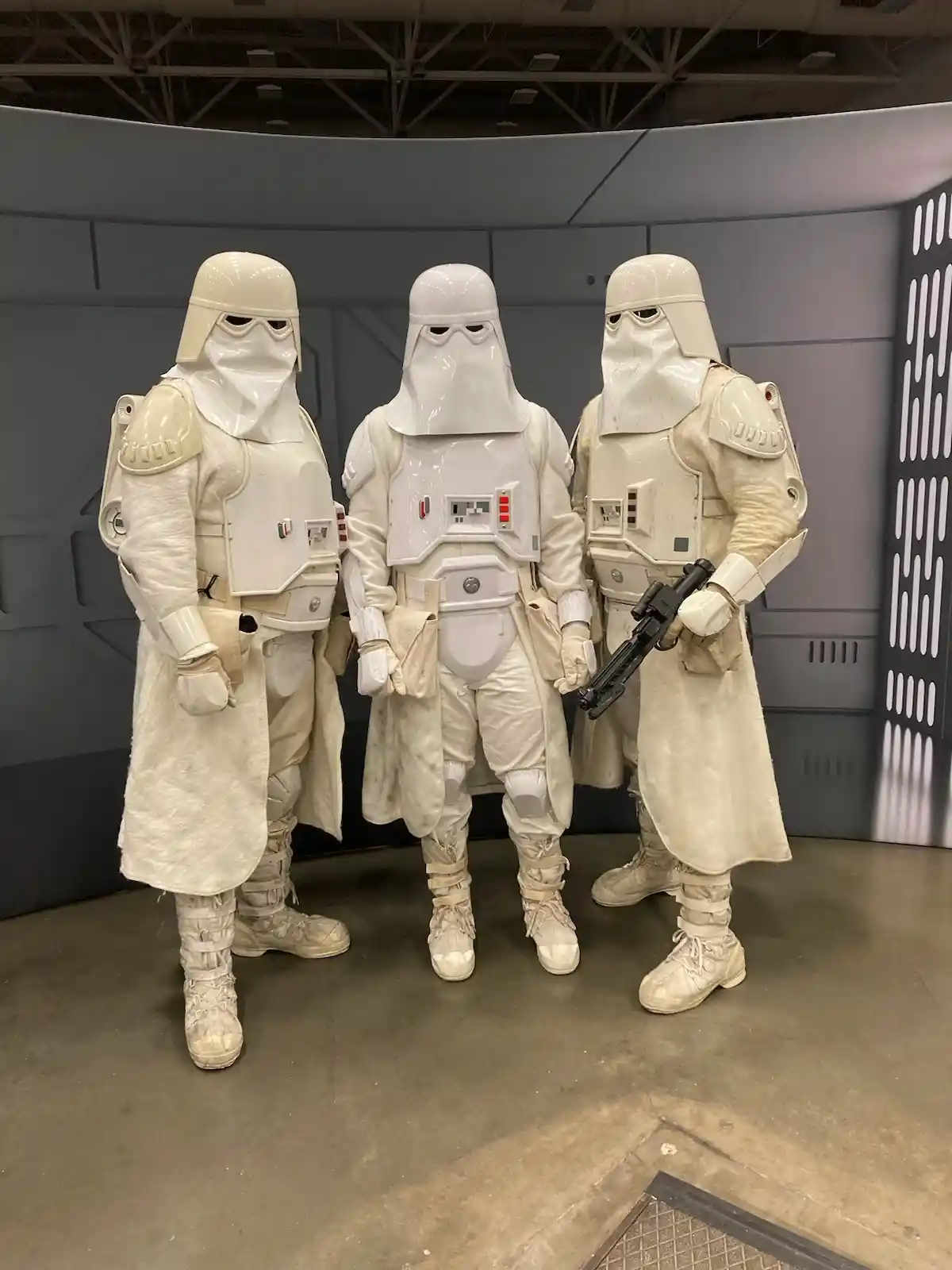 Three people in full white snowtrooper costumes from Star Wars stand side-by-side in a themed indoor setting. The central…