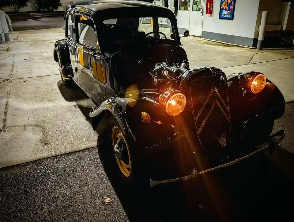A vintage black car with its hood open and headlights on is parked at a gas station at night. The car's engine is visible…