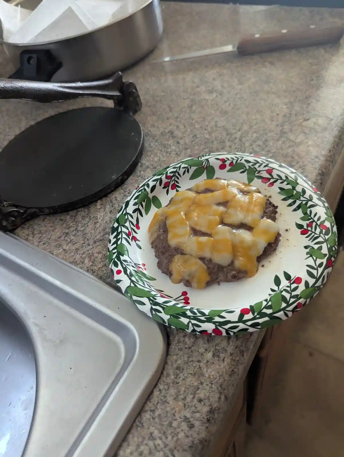A cooked hamburger patty topped with melted cheese sits on a paper plate with a green and red berry pattern, placed on a…