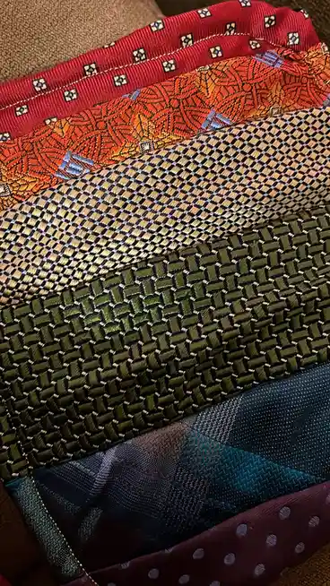 A close-up shot shows a stack of neatly folded, colorful neckties with various patterns including geometric, woven, and…