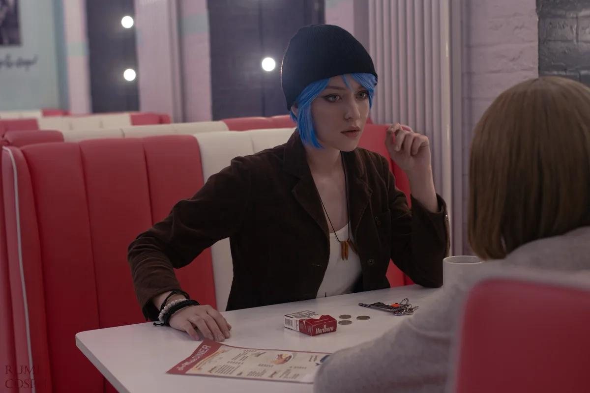 A person with blue hair and a black beanie sits at a diner booth, looking at another person across the table with a…