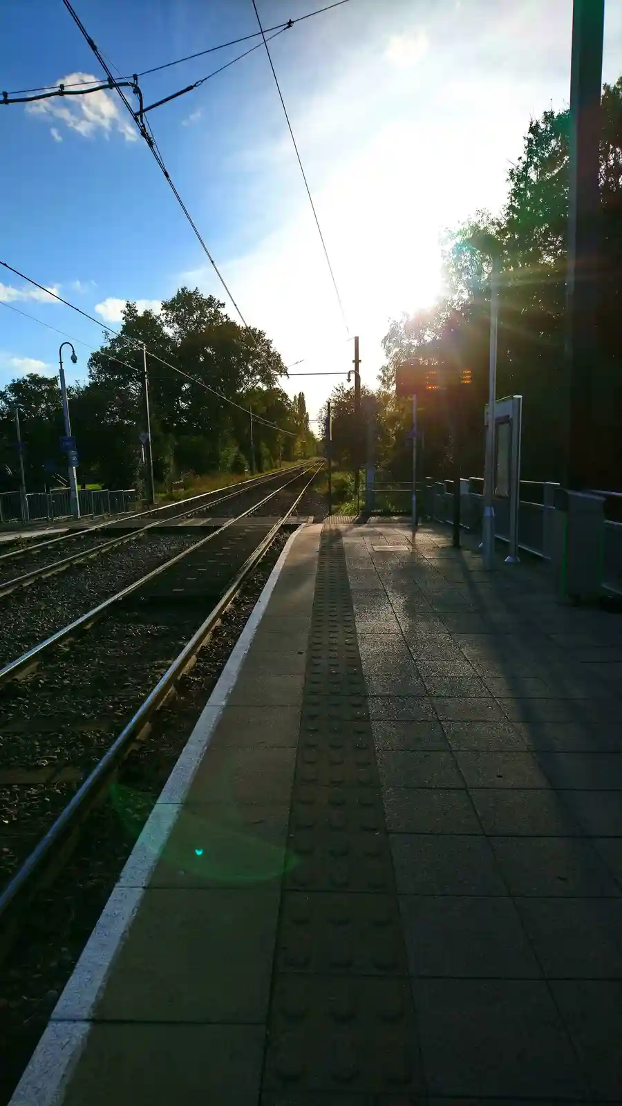 A photo of an empty train station platform at sunrise or sunset, with tracks receding into the distance under overhead wires…