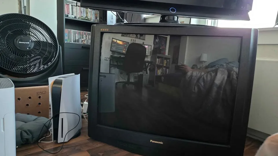 A black Panasonic CRT television sits on a wooden floor, its screen reflecting a room with a gaming setup, a fan, and a…