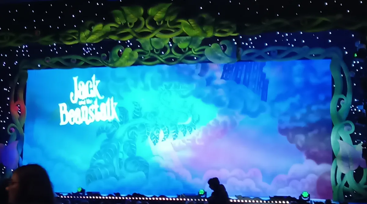 A stage show backdrop for 'Jack and the Beanstalk' features a large screen with the title in white cursive font against a…