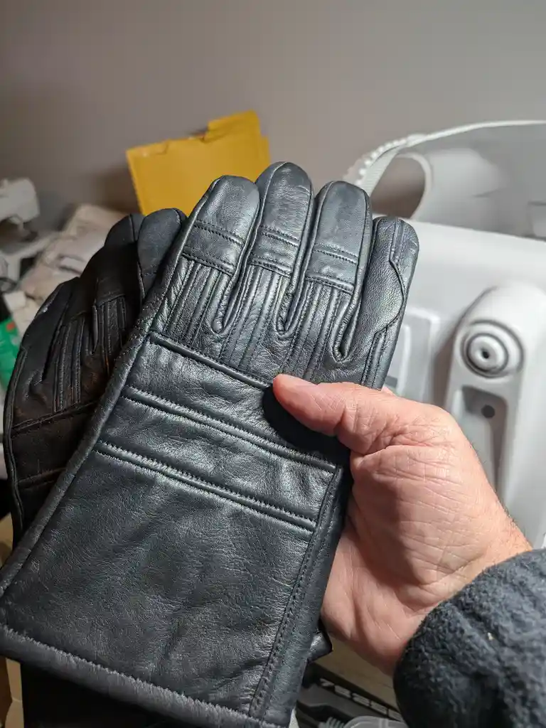 A person holds a pair of black leather gloves with detailed stitching, showcasing them to the camera. In the background, a…