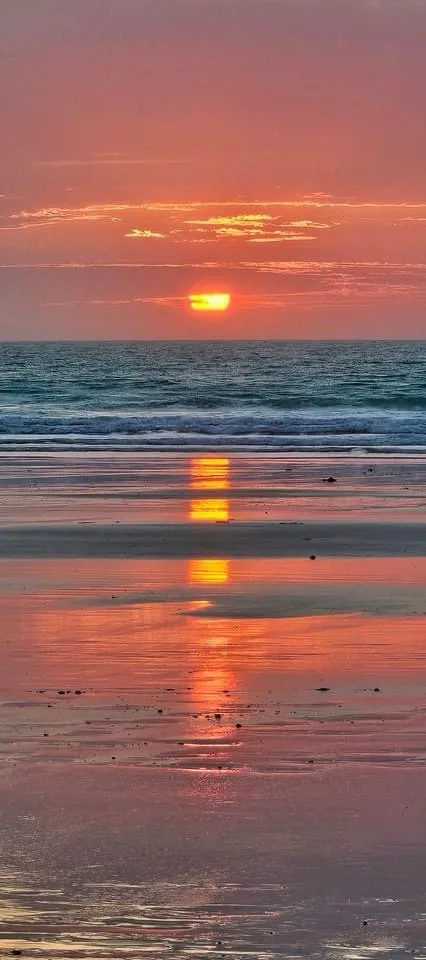 A serene beach sunset with the sun low on the horizon, casting orange and pink hues across the sky and reflecting vividly on…