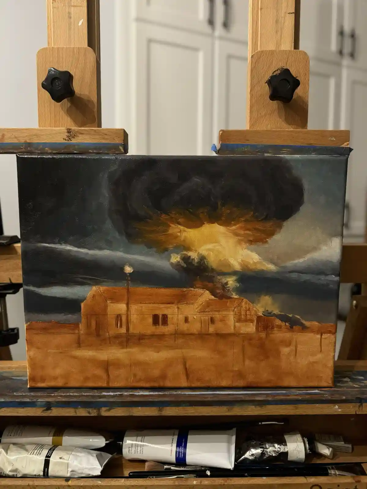 A painting on a canvas, mounted on an easel, depicts a large, stylized nuclear explosion above a simple building. The…
