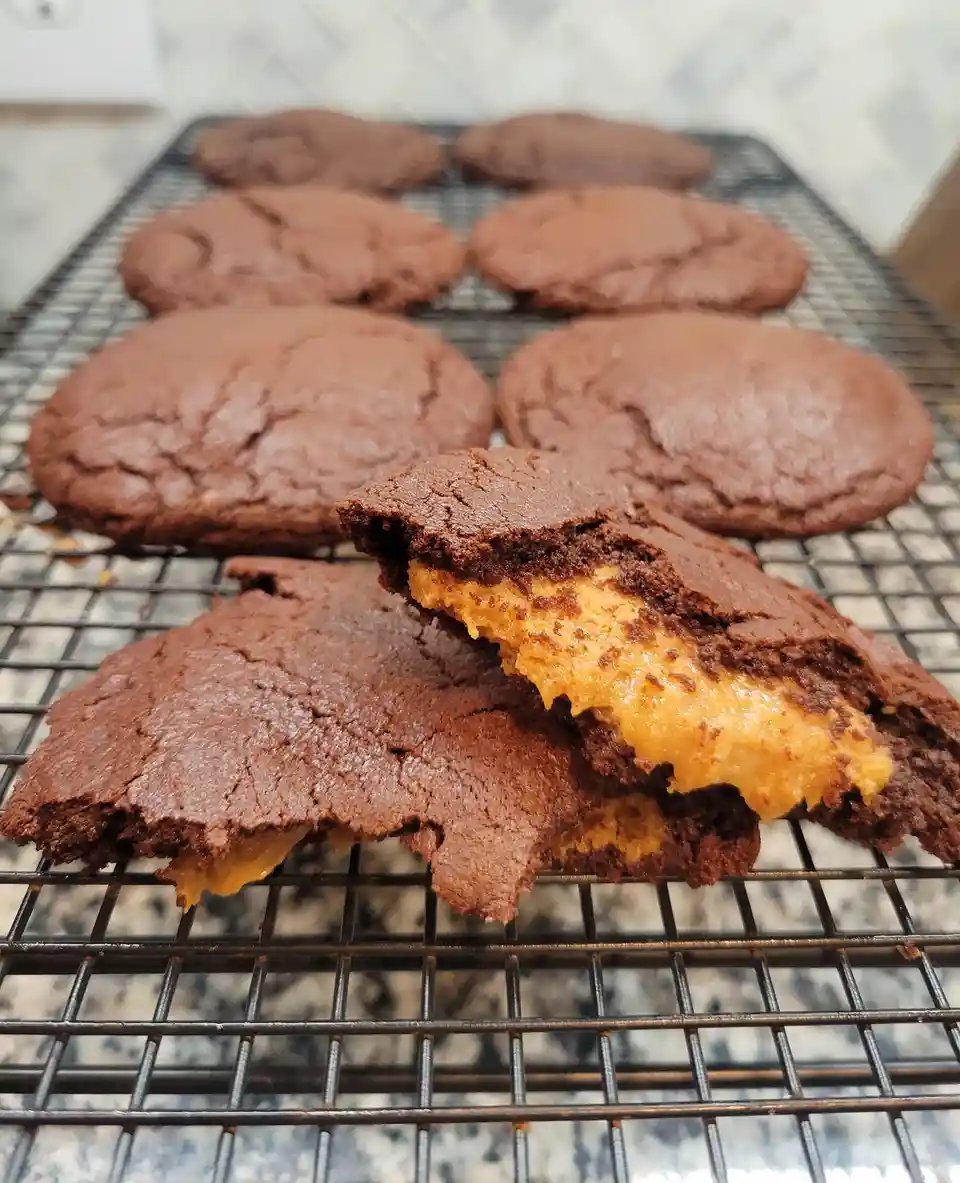 A close-up photo shows chocolate cookies on a wire cooling rack, with one broken open to reveal a gooey, orange-colored…