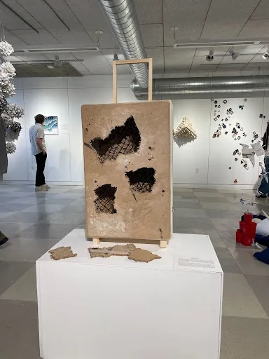 A large, beige, suitcase-shaped art installation with multiple black, net-like holes is displayed on a white pedestal in a…