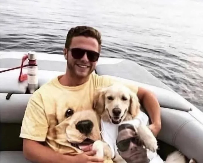 A smiling man wearing sunglasses sits in a boat holding two golden retrievers. One dog wears a white t-shirt with a photo of…