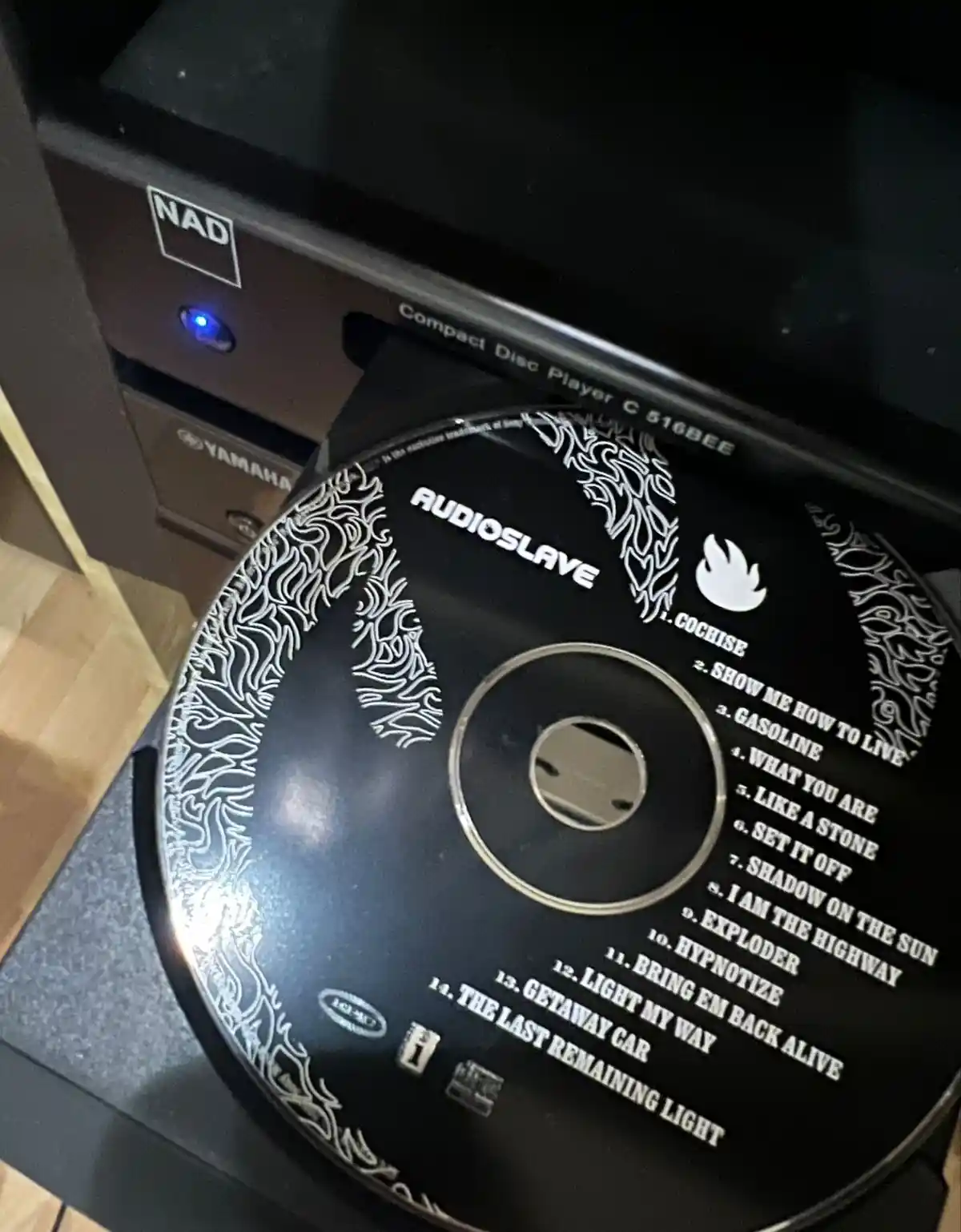 A black Audioslave CD with white text and flame-like border art is partially ejected from a NAD compact disc player. The…