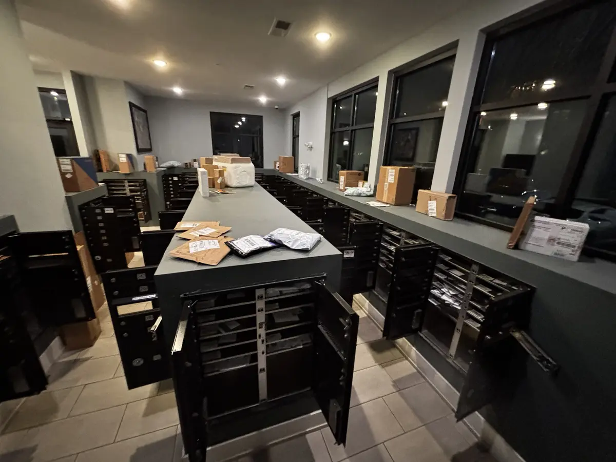 A long, dark gray counter in a mailroom is cluttered with packages and open mailboxes, with several large windows showing…