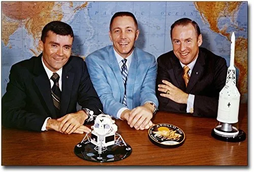 Three smiling men in suits sit at a wooden table in front of a world map, with a model of a lunar module, a circular…