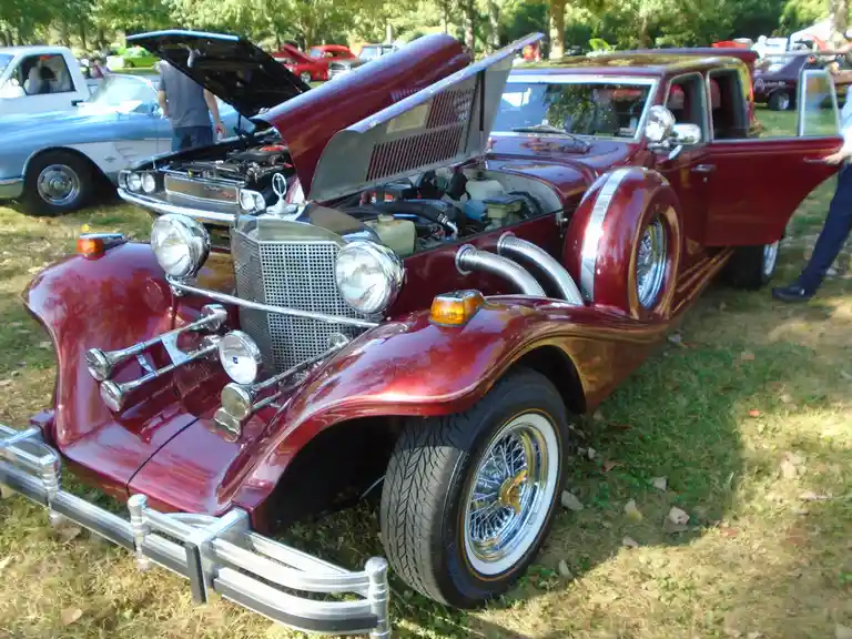 A shiny red custom classic car with its hood open, revealing a modified engine and chrome exhaust pipes, is parked on grass…