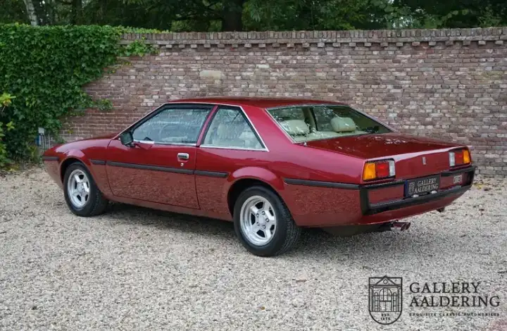 A maroon vintage two-door coupe is parked on gravel in front of a brick wall with ivy. The car has a sleek design with…