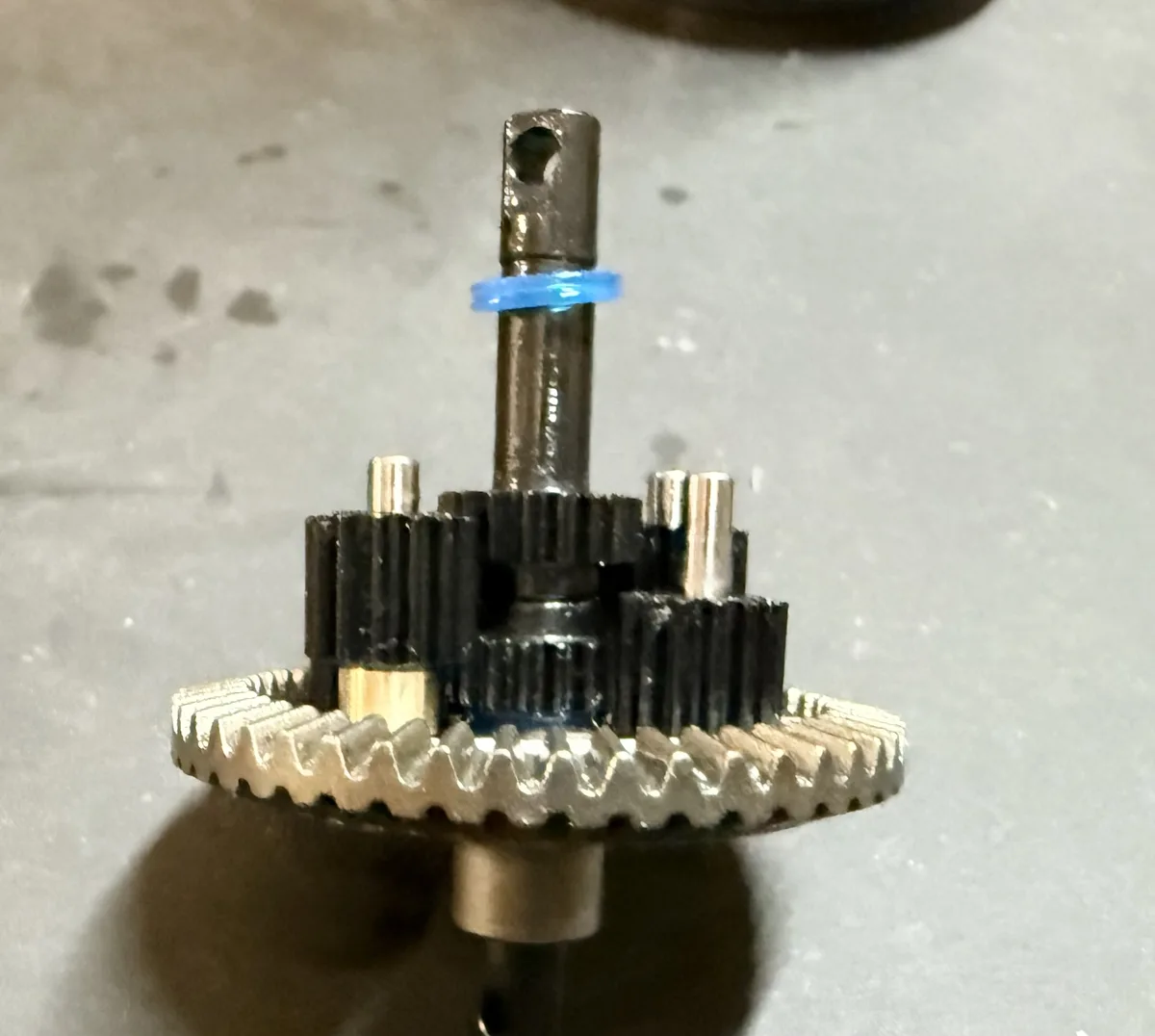 A close-up photo shows a mechanical gear assembly with a blue rubber band wrapped around its central shaft, holding it in…