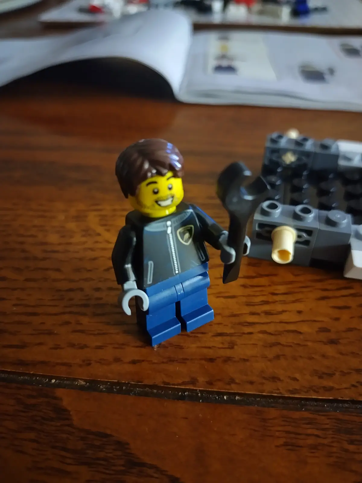 A smiling Lego minifigure with brown hair, wearing a black jacket with a yellow shield emblem and blue pants, stands on a…