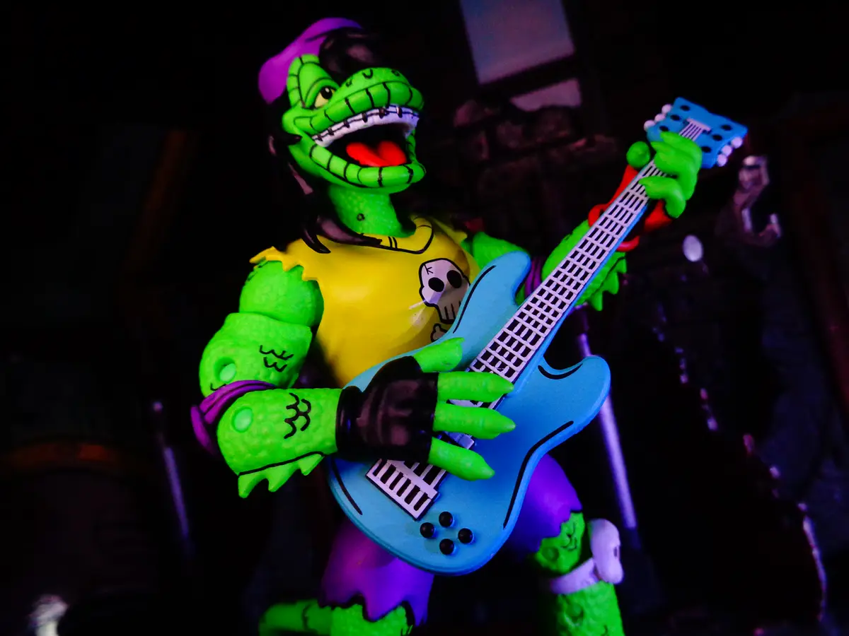 A green, cartoonish alligator-like action figure with a purple hat and yellow shirt is posed energetically holding a blue…