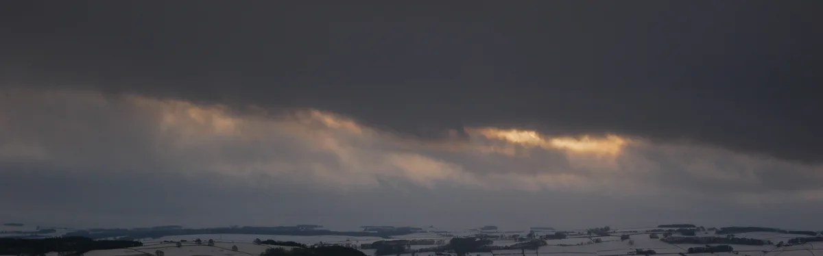 A wide landscape photo shows a snowy field under a dark, heavy cloud layer with a narrow band of golden sunlight breaking…