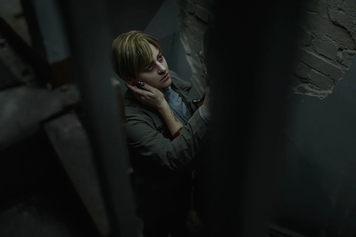 A person with short, light brown hair and dark nail polish is shown in a dimly lit, gritty environment, holding their own…