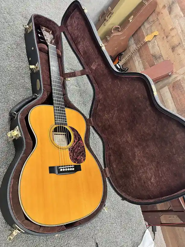 An acoustic guitar with a natural wood finish sits inside an open, empty black hardshell case lined with brown velvet…